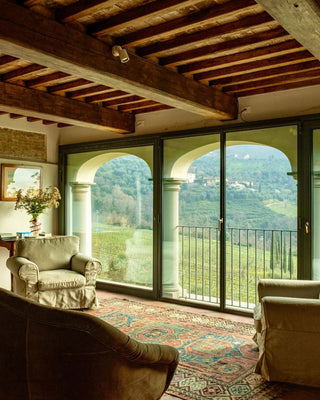 Soggiorno con travi in legno, divani e ampie vetrate ad arco affacciate sulle colline di Valgiano.