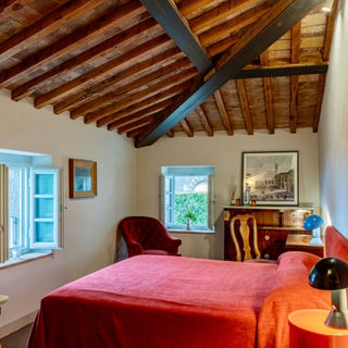 Camera matrimoniale di Villa Di Valgiano con copriletto rosso, scrittoio in legno e soffitto con travi a vista.