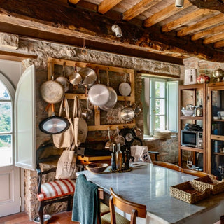 Cucina di Villa Di Valgiano con tavolo in marmo, pentole appese e pareti in pietra.