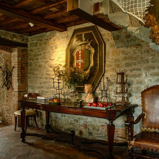 Tavolo in legno con candelabri e composizione floreale sotto uno stemma, nella sala da pranzo con pareti in pietra.